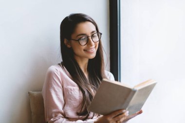 Boş zaman, eğitim, favori hobi konsepti. Neşeli kadın kafede en sevdiği kitabı okuyor, pencerenin yanında oturuyor, boş yer var. Mutlu bayan zevkle vakit geçiriyor.