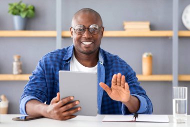 Çevrimiçi iletişim kavramı. Olgun siyah adam dijital tablet kullanıyor, selamlama hareketi yapıyor ve kameraya bakıyor, arkadaşlarına ya da ailesine video görüşmeleri yapıyor, karantina sırasında uzaktan sohbet ediyor.