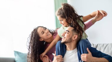 Üç kişilik neşeli bir Orta Doğulu ailenin evde birlikte eğlendiğinin portresi. Genç Arap ebeveynler küçük kızlarıyla oturma odasında oynuyorlar, anne, baba ve çocuk gülümsüyor ve gülüyorlar.