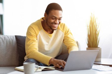 Memleketten online çalışan pozitif siyahi adam dizüstü bilgisayar kullanıyor, kopyalama alanı kullanıyor. Mutlu Afro-Amerikan genç adam oturma odasında kanepede otururken iş ortakları veya iş arkadaşlarıyla sohbet ediyor.