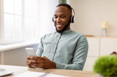 Neşeli Afrikalı iş adamı telefon kullanıyor, kulaklık takıyor, cep telefonu çalıyor ya da internetten iş yerinde mola vermiş müzik dinliyor modern ofiste oturuyor. Seçici Odaklanma