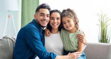 Evde üç poz veren mutlu, genç, orta doğulu bir ailenin portresi, anne, baba ve tatlı küçük kızları oturma odasında koltukta kucaklaşıyorlar ve kameraya gülümsüyorlar, birlikte vakit geçiriyorlar.