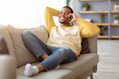 Neşeli Afro-Amerikan milenyum adamı oturma odasında kanepede dinleniyor, cep telefonuyla arkadaşlarıyla veya sevgilileriyle konuşuyor ve fotokopi odasına bakıyor, hafta sonunun keyfini çıkarıyor, ev içinin tadını çıkarıyor.