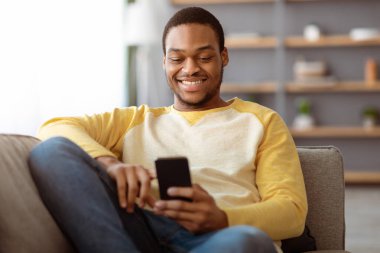 Evdeki kanepede uzanan neşeli siyahi adam, cep telefonu kullanıyor, arkadaşlarıyla sohbet ediyor ya da online oyunlar oynuyor, kopyalama alanı kullanıyor. Mutlu Afrikalı Amerikalı genç adam cep telefonunda sosyal medya kullanıyor.