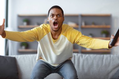Öfkeli siyahi adam TV 'de futbol maçı izliyor, elinde uzaktan kumandayla bağırıyor ve el sallıyor, Afro-Amerikan genç adam futbol takımını kaybetmeye sinirlendi, iç mekan, fotokopi...