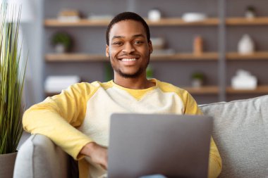 Evinde dizüstü bilgisayar kullanan neşeli siyahi adamın yakın çekimi. Yakışıklı Afro-Amerikan genç adam kanepede oturup internette sohbet ediyor, serbest çalışan, kameraya gülümseyen.