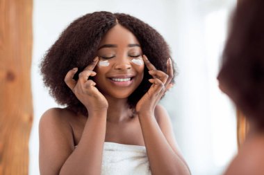 Büyüleyici bir Afro-Amerikan kadın. Evdeki aynanın yanında gözlerinin altında besleyici yüz kremi sürüyor. Güzel siyah kadın kendine iyi bakıyor, cildini besliyor.