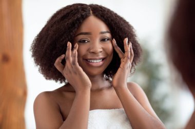 Güzel siyah bayan aynanın önünde duruyor, ilk kırışıklıkları kontrol ediyor, evde krem sürdükten sonra tenine dokunuyor. Cilt bakımı sonrası yansımasına bakan Afrikalı Amerikalı bir kadın.