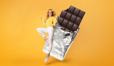 Sarı bluzlu ve beyaz pantolonlu neşeli bir kadın heyecanla atlıyor. Arkasında, parlak ambalajlı büyük bir çikolata, neşeli atmosfere neşe katıyor ve enerjik bir hava yaratıyor..