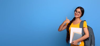 Mutlu Hintli kız öğrenci gözlük takıyor işaret işareti işareti yapıyor, kolej malzemeleri tutuyor ve sırt çantası takıyor, mor stüdyo arka planında tek başına duruyor bedava fotokopi alanı, afiş