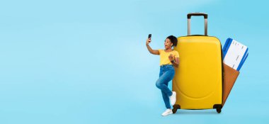 Bir gezgin, parlak sarı bir bavula yaslanırken akıllı telefonuyla etkileşime girer. Yaz tatilinin heyecanı seyahat planlarının ortaya çıkmasıyla ortaya çıkıyor. Bu an, rezervasyon gezilerinin neşesini yakalıyor..