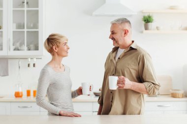 Günaydın, harika nazik ruh hali, taze kahve içkisi ve kahvaltıda evde beraberiz. Orta yaşlı mutlu bayan ve erkek, modern mutfağın içinde sıcak içeceklerle dolu bardaklarla, günışığında, boşluğu kopyalayın.