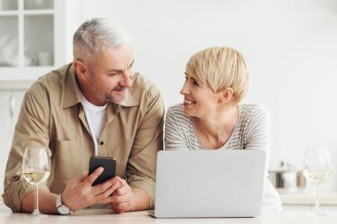 Teknoloji ve sosyal medyada boş zamanı olan orta yaşlı bir çift. Gülümseyen yetişkin kadın ve erkek, modern mutfağın içinde birbirlerine bakıyorlar. Meyveler, bir bardak şarap, dizüstü bilgisayar ve telefonla.
