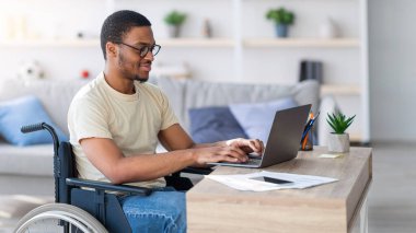 Tekerlekli sandalyedeki siyahi adam internetten bilgisayar kullanıyor, internette iş arıyor. Özürlü genç bir adam iş görüşmesi yapıyor, internette iş projesi yapıyor.