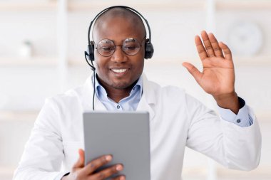 Modern dijital teknolojiler ve terapistle istişare. Mutlu Afro-Amerikan doktor tablete el sallıyor, video görüşmesi yapıyor, hastanedeki kabinede oturuyor.