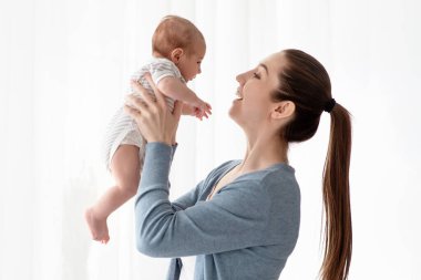 Doğum İzni Konsepti. Mutlu genç bir kadın, yeni doğmuş çocuğuyla evde vakit geçiriyor, Millennial bir anne, küçük oğluyla oynuyor ve onu havaya kaldırıyor, anneliğin tadını çıkarıyor, yandan bakıyor.