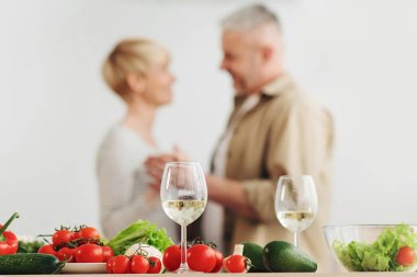 Romantik randevu, aile ilişkileri. Minimalist beyaz mutfak içinde dans eden mutlu yaşlı çift. Şaraplı bardaklara odaklanın ve parlak sebzeler ve masada salata, bulanık