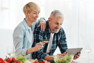Çevrimiçi alışveriş ve market alışverişi. Gülümseyen orta yaşlı aile tablete bakar ve mutfak içinde parlak sebzeler ve şarap kadehleriyle kredi kartı tutar.