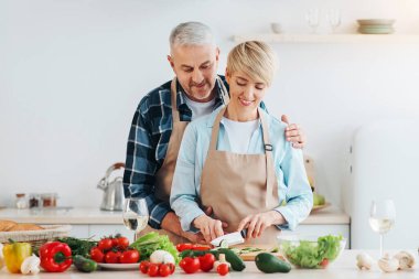 Sağlıklı bakım, yararlı yaşam tarzı, düzgün yemek, vejetaryen ve ev yapımı yemek. Gülümseyen olgun bir kadın yemek pişiriyor, erkek mutfakta kadına sarılıyor. Parlak sebzelerle, masada bir kadeh şarapla, boş bir alanda.