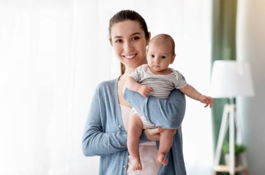 Mutlu Anne Sevgisi. Gülümseyen güzel bir kadının portresi, yeni doğmuş bebeğiyle evde poz veren neşeli bir annenin sevimli bebeğini kucağında tutması, anneliğin tadını çıkarması.