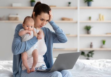 Çalışan anne sorunları. Bebeğini kucağında tutan ve dizüstü bilgisayar ekranına bakan genç bir annenin evde çocuğuna bakarken uzak bir işte sorun yaşamasından endişe duyması.