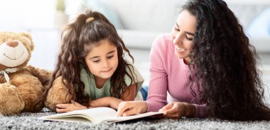 Çocuk Geliştirme. Genç anne, küçük kızına kitap okurken birlikte oturma odasında dinleniyorlar, anne ve çocuk eğleniyor ve evde eğleniyorlar, panorama...