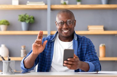Akıllı telefon kullanan, kameraya bakıp gülümseyen, karantina sırasında uzaktan sohbet eden neşeli, olgun Afrikalı Amerikalı adam.