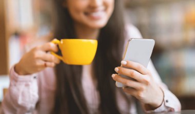 Kahve molası. Kafede akıllı telefon kullanan ve sıcak içecek içen mutlu Arap kadın, yakın plan. Kafeteryada bir fincan kahve ve cep telefonuyla dinlenen Latin bir kadın.