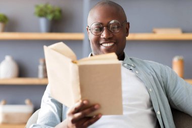 Boş zaman konsepti. Siyahi olgun adam kitap okuyor, uzun kurgu romanlarının tadını çıkarıyor, seçici odaklanma. Afrika kökenli Amerikalı okuyucu gözlük takıyor ve hafta sonu evde dinleniyor..