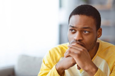 Sorunlu siyahi adam bir şey düşünürken fotokopi odasına bakıyor. Gri oturma odasındaki kanepede oturuyor, yakın plan yapıyor. Üzgün Afrikalı Amerikalı genç adam ellerine yaslanıyor, sorun yaşıyor.