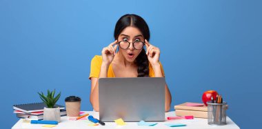 Omg, inanılmazsın. Şaşırmış gözlüklü Hintli kadının portresi masada dizüstü bilgisayar kullanarak mor stüdyo arka planında izole edilmiş bilgisayar ekranına bakıyor. Bayan şok edici haberler okuyor.