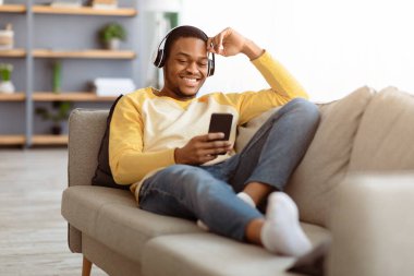 Mutlu siyah adam evdeki koltukta dinlenirken cep telefonundan video izliyor, kablosuz kulaklık kullanıyor, kopyalama alanı kullanıyor. Neşeli Afrikalı Amerikalı genç adam akıllı telefondan film izliyor.