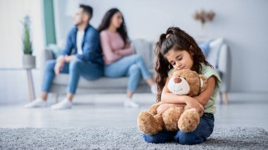 Çocuklar ve Aile Çatışmaları Konsepti. Oturan küçük bir kızın portresi, ebeveynleri evde tartıştıktan sonra üzgün, üzgün çocuk oyuncak ayıya sarılıyor, depresif ve yalnız hissediyor, seçici bir şekilde çocuğa odaklanıyor.