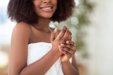 Duştan ya da banyodan sonra el kremi süren, beyaz havlu takan, evcil cilt bakımının keyfini çıkaran, vücudunu kaplayan genç siyahi bir kadın görüntüsü. Güzellik ve refah kavramı