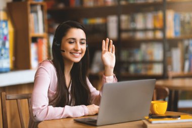 Kafede dinlenirken görüntülü konuşma yapan heyecanlı Latin bayan dizüstü bilgisayar, kulaklık ve fotokopi odası kullanıyor. Mutlu kadın dizüstü bilgisayara el sallıyor, video konferansına katılıyor ve kahve içiyor.