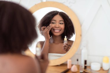 Güzellik rutin prosedürleri. Şehvetli genç siyahi kadın evdeki aynanın önüne su serumu sürüyor. Çekici Afrikalı Amerikalı kadın makyaj masasında kozmetik ürünler kullanıyor.