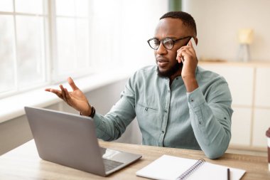 Ciddi bir siyahi işadamı cep telefonuyla konuşuyor. İş yerindeki dizüstü bilgisayarın başında. Modern ofiste masasında oturuyor. İş İletişim Konsepti. Seçici odak