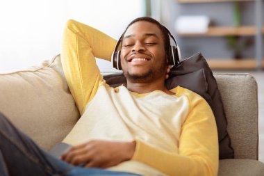 Kablosuz kulaklıklı mutlu siyahi adamın yakın plan portresi. Kanepede yatar ve evde müzik dinlerken gülümserken, elinde akıllı telefon tutarken. Mutlu Afrikalı Amerikalı adam yeni müzik kulaklıklarının keyfini çıkarıyor.