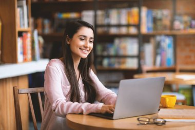 Uzaktan öğrenme ve online çalışma konsepti. Mutlu Arap kadın rahat şehir kafesinde dizüstü bilgisayar kullanıyor, boş yer var. Milenyum Latin bayanı serbest çalışıyor, web konferansına katılıyor.