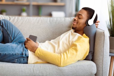 Rahat Afro-Amerikalı adam evdeki koltukta dinlenirken cep telefonuyla müzik dinliyor, kablosuz kulaklık kullanıyor, fotokopi çekiyor. Akıllı telefonda müzik uygulaması kullanan neşeli siyahi adam.