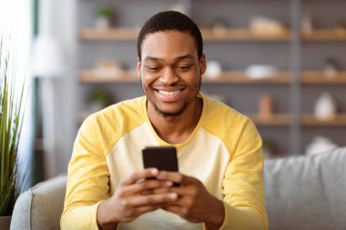 Afro-Amerikalı bir adam akıllı telefondan sevgilisiyle sohbet ediyor ve evde dinlenirken gülümsüyor, yakın plan portre, fotokopi odası. Mutlu siyah adam cep telefonundaki güzel flört uygulaması ile eğleniyor.