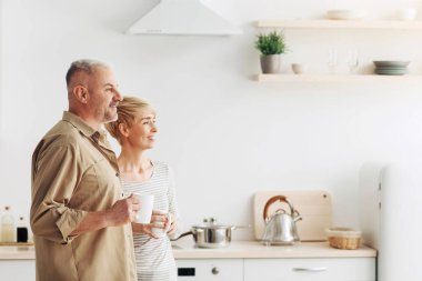 Harika bir teklif, reklam, birlikte kahvaltı, sabah kahvesi, nazik bir ruh hali. Gülümseyen orta yaşlı bir çift sıcak bir bardak içecekle mutfaktaki boş yere bakıyor.