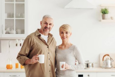 Pozitif duygular, neşeli ruh hali, günaydın, kahve molası, kahvaltı konforu ve ev konforu. Minimalist mutfak iç mekanında bir fincan sıcak içecekle gülümseyen yetişkin Avrupalı çift.