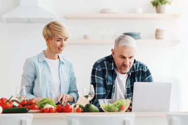 İnternette yemek tarifi ara, yemek pişir ve blog takipçileriyle iletişim kur. Konsantre orta yaşlı adam dizüstü bilgisayarda çalışıyor, kadın yemek hazırlıyor, masada sebzeler ve modern mutfak içleri var.