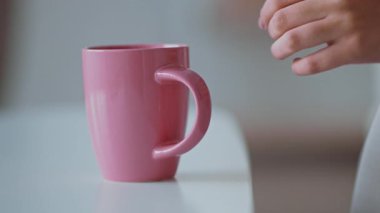 Genç bir kadın evdeki masada duran pembe kupaya uzanıyor. Sıcaklıkla dolu rahat bir hayat ortamında rahat bir anın tadını çıkarıyor..