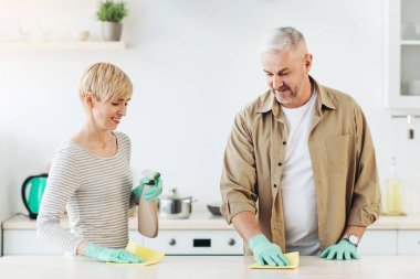 Gülümseyen kadın ve adam deterjan spreyiyle mutfak masasından bez parçasıyla toz temizliyor. Yetişkin bir çift mavi eldivenli modern beyaz konforlu apartman dairesini temizliyor.