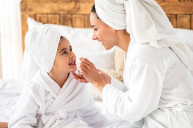 Anne küçük tatlı kızına makyaj yapıyor, yan görünüm, yakın çekim. Bornoz giymiş, saçı havluya sarılı, çocuk dudaklarına ruj sürmüş, yatak odasının iç tarafına...