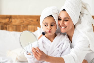 Güzel anne, küçük kızının yanağına allık sürüyor, aynaya bakıyor ve gülümsüyor, yakın plan, kopyalama alanı. Neşeli anne ve kız bornoz giyer, yatağa oturur, makyaj malzemelerinin tadını çıkarırlar.