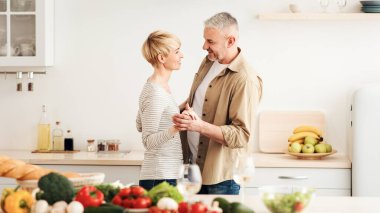 Eğlence, mutlu ve birlikte çıkma, Covid-19 salgını sırasında orta yaşlı ilişkiler. Neşeli son sınıf çifti, rahat mutfağın içinde dans ediyor. Parlak sebzeler, şarap kadehleri, boş alanlar.