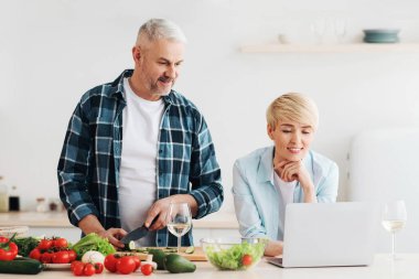 Yemek blogcusu, serbest çalışan, internetten yemek tarifleri yapan ve birlikte yemek yapan. Dost canlısı orta yaşlı koca sebzeleri kesiyor, karısı mutfakta dizüstü bilgisayara bakıyor ve masasında parlak sebzeler var.